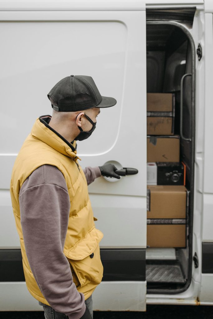 A courier in a yellow vest delivers packages from a van, emphasizing the new normal with safety protocols.