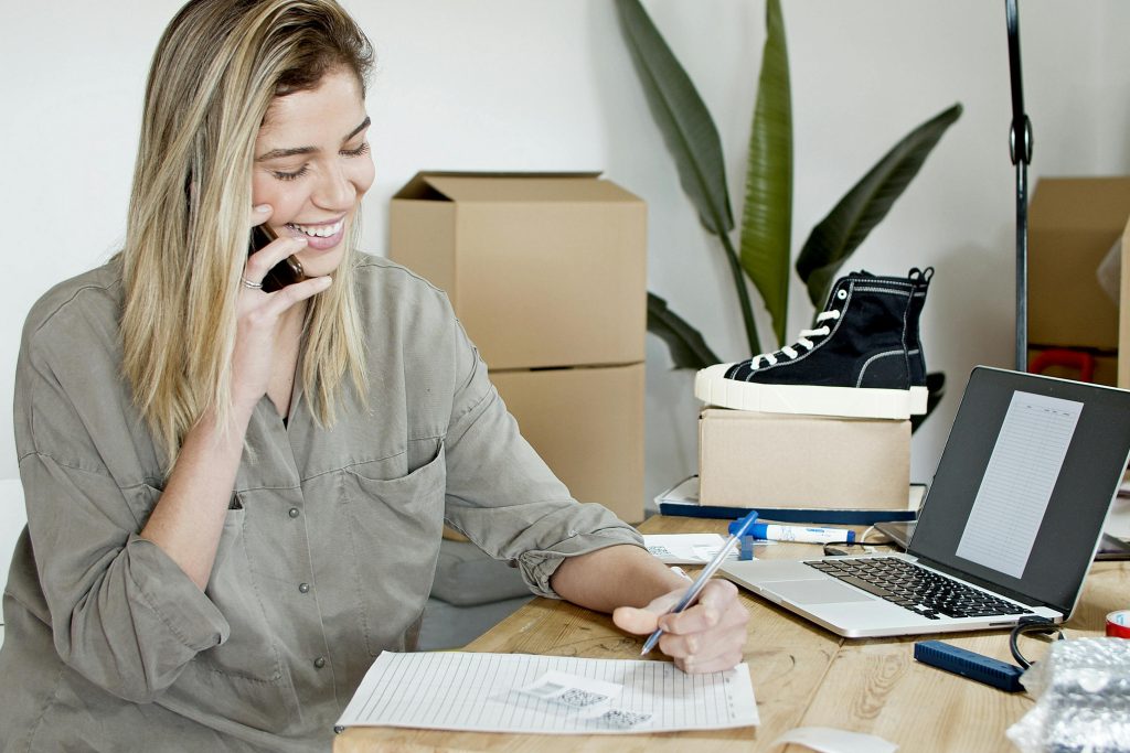 A happy businesswoman multitasks with a laptop, phone, and notes at her office desk, managing her online store orders.